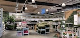 A well-organized store section filled with a diverse array of potted plants and flowers. The display includes tall green plants, colorful flowers, and various sizes of plant pots. Shelves along the walls hold additional plants and decorative greenery. Overhead, industrial lighting fixtures are visible, highlighting the vibrant colors of the plants below.