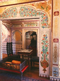 An ornately decorated room features vivid, intricate floral patterns on the walls, complemented by gold accents. The room contains a chess table with pieces set up, surrounded by dark wooden chairs. A wooden daybed with orange cushions is present in the foreground. The overall decor suggests a rich, cultural ambiance.