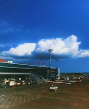 A modern airport terminal with a clear blue sky and large fluffy clouds overhead. The foreground features a tarmac with various ground service vehicles, including a small truck with a person driving it. The terminal building has several large glass windows and is surrounded by lighting poles.