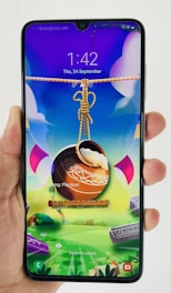 A hand holds a smartphone displaying a colorful and vibrant lock screen. The screen features a digital artwork with a pot suspended by a rope, accompanied by a flute. The background is a whimsical landscape with bright colors and abstract shapes.