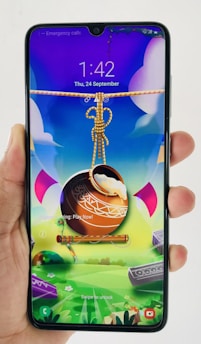 A hand holds a smartphone displaying a colorful and vibrant lock screen. The screen features a digital artwork with a pot suspended by a rope, accompanied by a flute. The background is a whimsical landscape with bright colors and abstract shapes.