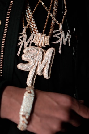 Several chains featuring large, glittering pendants and a bracelet are showcased, all encrusted with sparkling stones. The pendants and the bracelet have a gold hue, with the letters 'SM' prominently displayed on some of the pendants.