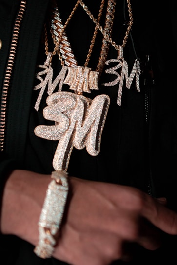 Several chains featuring large, glittering pendants and a bracelet are showcased, all encrusted with sparkling stones. The pendants and the bracelet have a gold hue, with the letters 'SM' prominently displayed on some of the pendants.