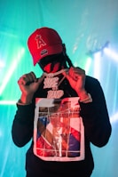 A person wearing a red cap and a graphic sweatshirt is holding a large, glittery chain with prominent lettering. The background is illuminated with teal and blue lights, creating an atmospheric ambiance.