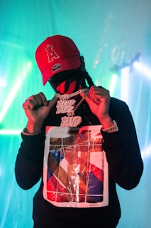 A person wearing a red cap and a graphic sweatshirt is holding a large, glittery chain with prominent lettering. The background is illuminated with teal and blue lights, creating an atmospheric ambiance.