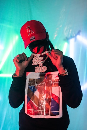 A person wearing a red cap and a graphic sweatshirt is holding a large, glittery chain with prominent lettering. The background is illuminated with teal and blue lights, creating an atmospheric ambiance.