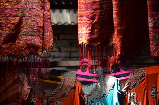 Colorful woven textiles hang on a wooden rod under natural light. The fabric displays intricate patterns in vibrant red, orange, and blue hues. Below, clothes with detailed embroidery are visible on pink and yellow hangers. A brick wall is partially visible in the background, adding to the rustic feel.