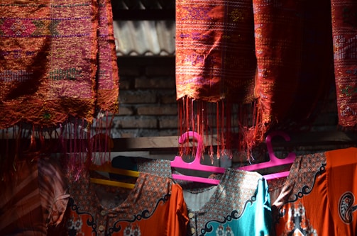 Colorful woven textiles hang on a wooden rod under natural light. The fabric displays intricate patterns in vibrant red, orange, and blue hues. Below, clothes with detailed embroidery are visible on pink and yellow hangers. A brick wall is partially visible in the background, adding to the rustic feel.