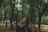 A person stands with eyes closed in a serene forest setting. Trees with lush green foliage surround the individual, creating a peaceful and contemplative atmosphere.