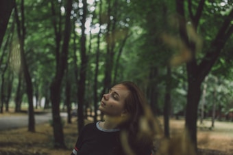 A person stands with eyes closed in a serene forest setting. Trees with lush green foliage surround the individual, creating a peaceful and contemplative atmosphere.