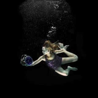 A woman is underwater, gracefully holding a large alarm clock. She is dressed in a dark, shimmering outfit and her hair is flowing freely, creating a surreal scene. Bubbles rise above her as she navigates the dark water.