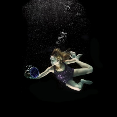 A woman is underwater, gracefully holding a large alarm clock. She is dressed in a dark, shimmering outfit and her hair is flowing freely, creating a surreal scene. Bubbles rise above her as she navigates the dark water.