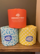 Three rolls of toilet paper with colorful wrappers are placed on a wooden shelf. The top roll has an orange wrapper labeled 'EMERGENCY ROLL!!' with humorous text. The two rolls at the bottom have blue and yellow wrappers with abstract patterns and the brand name 'who gives a crap'. The setting appears to be a bathroom with a neutral wall background.