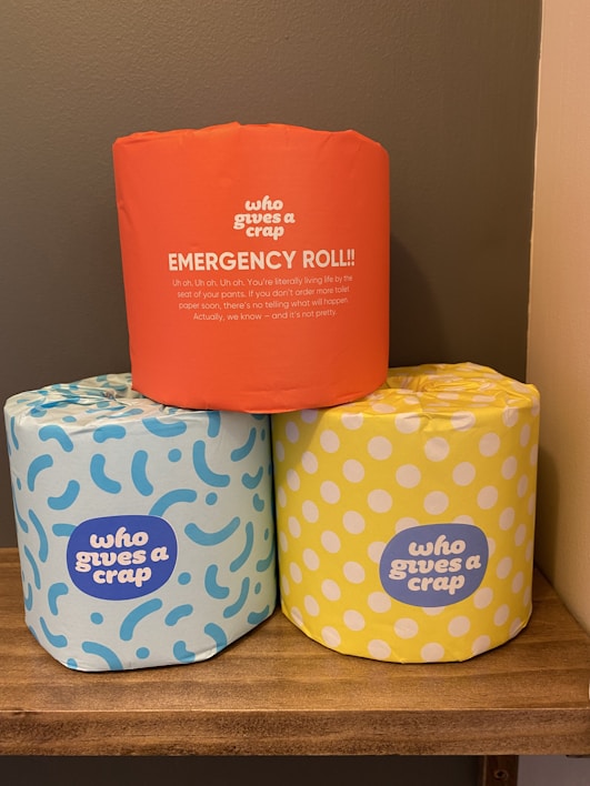 Three rolls of toilet paper with colorful wrappers are placed on a wooden shelf. The top roll has an orange wrapper labeled 'EMERGENCY ROLL!!' with humorous text. The two rolls at the bottom have blue and yellow wrappers with abstract patterns and the brand name 'who gives a crap'. The setting appears to be a bathroom with a neutral wall background.