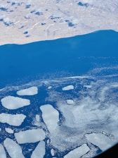 Aerial view of a body of water partially covered with floating ice patches. The landscape includes a coastline with a partially cloudy, rocky terrain, and vast open blue water.