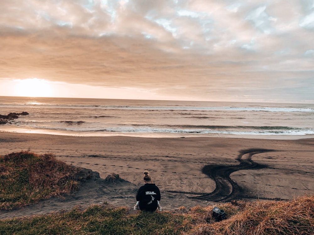 Persona con chaqueta negra sentada en roca marrón cerca del mar durante el día