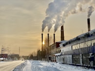 Four large industrial smokestacks are emitting significant amounts of smoke into the sky, with the sun partially visible through the smoke. The scene is set in a snowy environment with buildings and roads covered in snow. The sky is overcast with a yellowish tint.