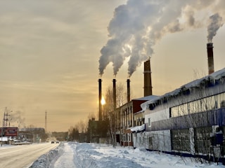 Four large industrial smokestacks are emitting significant amounts of smoke into the sky, with the sun partially visible through the smoke. The scene is set in a snowy environment with buildings and roads covered in snow. The sky is overcast with a yellowish tint.