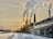 Four large industrial smokestacks are emitting significant amounts of smoke into the sky, with the sun partially visible through the smoke. The scene is set in a snowy environment with buildings and roads covered in snow. The sky is overcast with a yellowish tint.