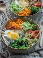 A bowl of colorful bibimbap featuring a sunny-side-up egg combined with a variety of vegetables including spiralized zucchini, carrots, sautéed spinach, and bean sprouts. There are also portions of kimchi and scattered sesame seeds on top. The arrangement is visually appealing with vibrant colors and textures.