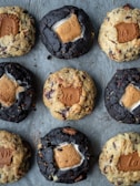 An arrangement of nine cookies on a baking sheet. The cookies are of two varieties: some are darker and others are lighter in color. Each cookie has a distinct filling of a marshmallow and a rectangular biscuit piece pressed into the center.