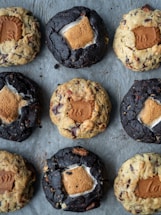 An arrangement of nine cookies on a baking sheet. The cookies are of two varieties: some are darker and others are lighter in color. Each cookie has a distinct filling of a marshmallow and a rectangular biscuit piece pressed into the center.