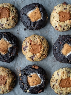 An arrangement of nine cookies on a baking sheet. The cookies are of two varieties: some are darker and others are lighter in color. Each cookie has a distinct filling of a marshmallow and a rectangular biscuit piece pressed into the center.