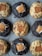 An arrangement of nine cookies on a baking sheet. The cookies are of two varieties: some are darker and others are lighter in color. Each cookie has a distinct filling of a marshmallow and a rectangular biscuit piece pressed into the center.