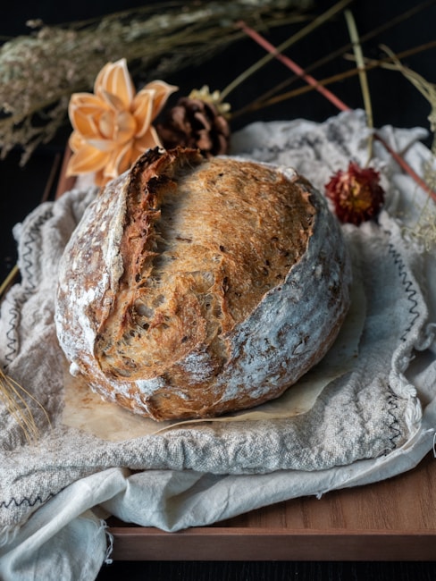 A rustic loaf of bread with a golden-brown crust rests on a cloth, surrounded by dried flowers and stems. The background is dark, highlighting the texture of the bread and the details of the floral arrangement.