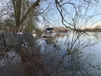 A flooded area where water has overflowed onto a path lined with a metal fence and large trees. A boat is moored nearby, surrounded by water and bare tree branches. The sky is clear, creating reflections in the water.