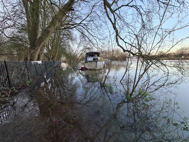 A flooded area where water has overflowed onto a path lined with a metal fence and large trees. A boat is moored nearby, surrounded by water and bare tree branches. The sky is clear, creating reflections in the water.