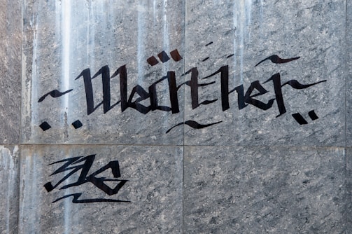 Graffiti art features stylized black lettering on a gray stone or concrete wall. The text is accompanied by abstract designs, with dynamic lines and flourishes.