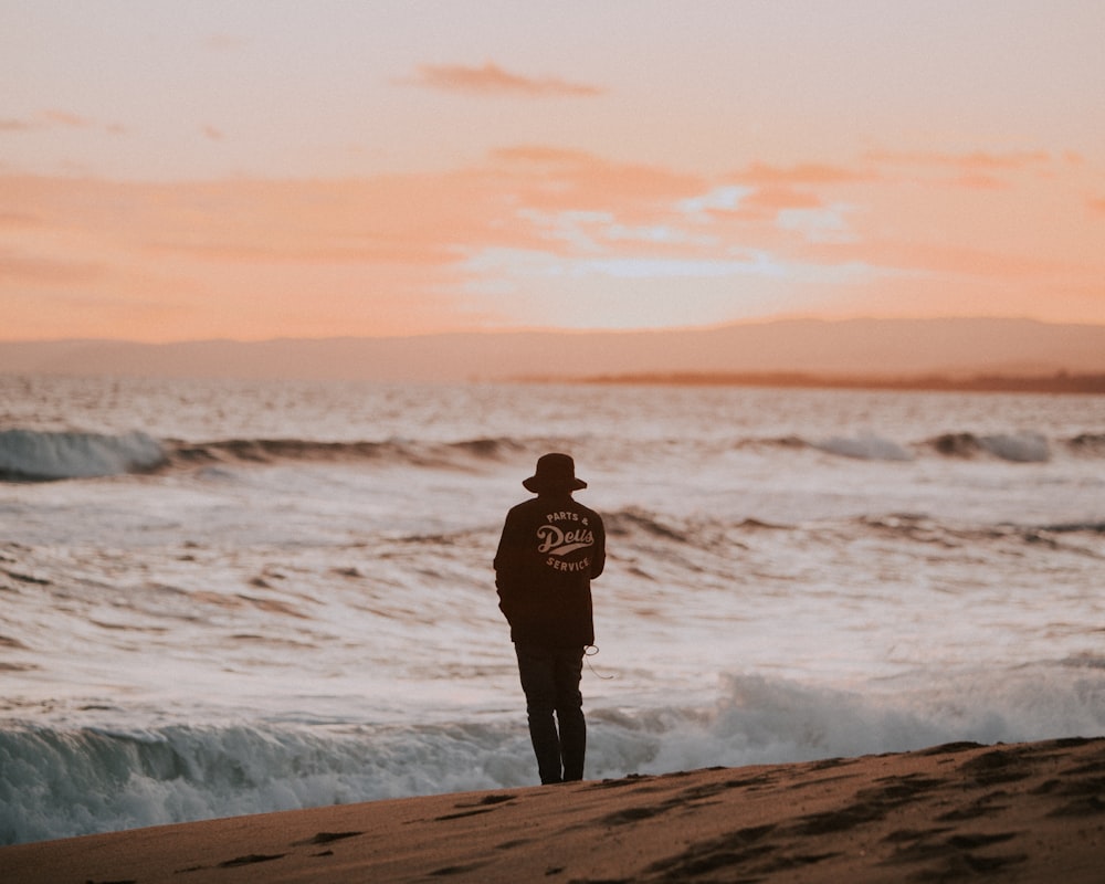 man in black jacket standing on seashore during sunset