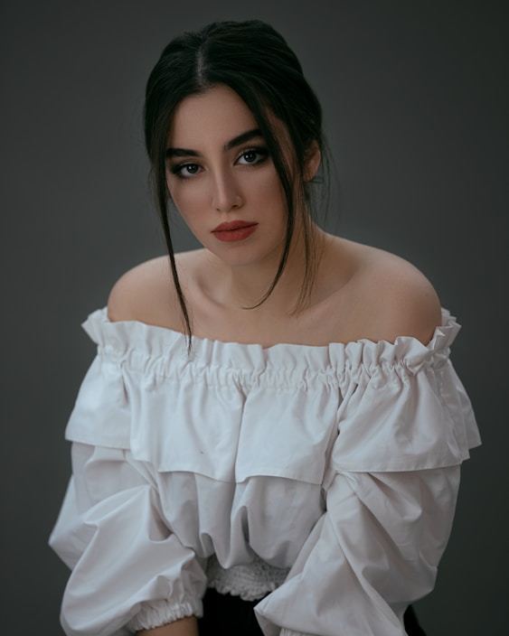A woman with long dark hair is wearing an off-the-shoulder white top with ruffled details. Her expression is neutral and she is looking directly at the camera. The background is a muted dark color, adding to the focus on her face and attire.
