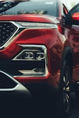 A close-up of a red car's front end, highlighting the sleek design of the headlight assembly and the smooth metallic finish of the body. The geometric shape of the headlights, chrome accents, and reflective surfaces add a sense of luxury and modernity.