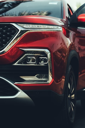 A close-up of a red car's front end, highlighting the sleek design of the headlight assembly and the smooth metallic finish of the body. The geometric shape of the headlights, chrome accents, and reflective surfaces add a sense of luxury and modernity.