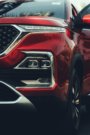 A close-up of a red car's front end, highlighting the sleek design of the headlight assembly and the smooth metallic finish of the body. The geometric shape of the headlights, chrome accents, and reflective surfaces add a sense of luxury and modernity.