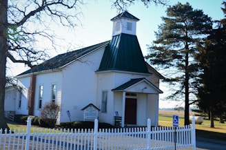 A small, white church with a green metal roof, set in a rural area with open fields in the background. The church has a steeple and a red door, surrounded by a white picket fence. There are leafless trees nearby, and a handicapped parking sign is visible.