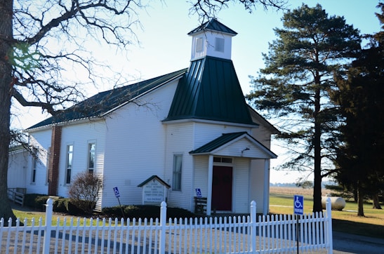 A small, white church with a green metal roof, set in a rural area with open fields in the background. The church has a steeple and a red door, surrounded by a white picket fence. There are leafless trees nearby, and a handicapped parking sign is visible.