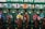 A group of jockeys and horses are lined up in starting gates at a horse racing track. The gate shows numbers from 5 to 9, and each horse is equipped with colorful saddle blankets and headgear. The jockeys are wearing brightly colored uniforms and helmets. The setting indicates the beginning of a horse race.