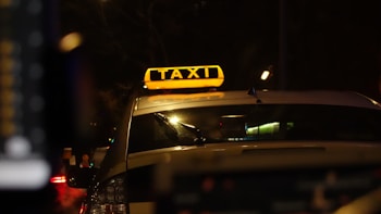 A taxi with a bright yellow sign on top, illuminated against the backdrop of a dark street. The surroundings appear to be urban, with the blurred lights of traffic visible in the background. The angle is from behind the vehicle, focusing on the signage.
