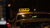A taxi with a bright yellow sign on top, illuminated against the backdrop of a dark street. The surroundings appear to be urban, with the blurred lights of traffic visible in the background. The angle is from behind the vehicle, focusing on the signage.