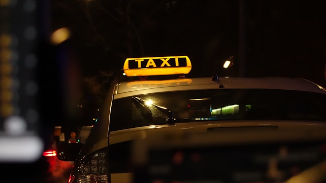 A taxi with a bright yellow sign on top, illuminated against the backdrop of a dark street. The surroundings appear to be urban, with the blurred lights of traffic visible in the background. The angle is from behind the vehicle, focusing on the signage.