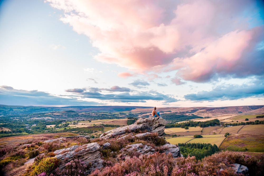 person in green shirt sitting on rock near green grass field under white clouds and blue