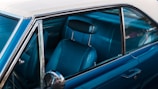 The image captures the interior of a vintage car, focusing on the front passenger seat and the window. The upholstery is made of blue leather with a ribbed texture, and the window is partially open, showing a reflective silver side mirror. The exterior paint is a glossy blue, and the roof of the car is a distinctive off-white shade.