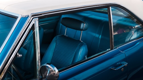 The image captures the interior of a vintage car, focusing on the front passenger seat and the window. The upholstery is made of blue leather with a ribbed texture, and the window is partially open, showing a reflective silver side mirror. The exterior paint is a glossy blue, and the roof of the car is a distinctive off-white shade.