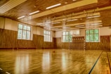 A spacious indoor gymnasium with a wooden floor, featuring a basketball hoop and a goalpost. The room is well-lit with large windows allowing natural light to come in. Gymnastic rings hang from the ceiling, and benches are placed along the walls.