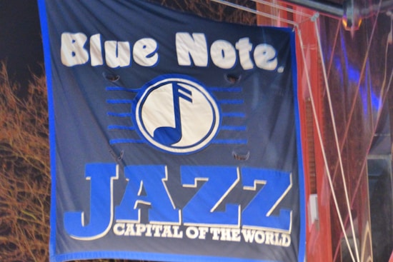 A dark blue banner featuring the text 'Blue Note' and 'JAZZ CAPITAL OF THE WORLD' in bold white and blue letters. In the center, a stylized music note design is enclosed in a circle.