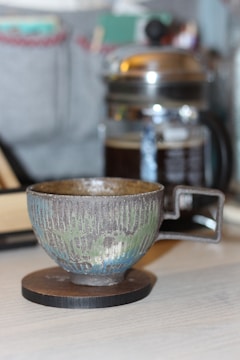 A ceramic cup with a textured surface featuring green and brown hues stands on a round wooden coaster. In the background, there is a French press filled with dark coffee. The setting appears casual and cozy, with some out-of-focus books or objects adding to the background ambiance.