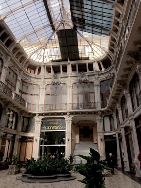 An elegant interior space featuring a high glass ceiling with ornate metalwork and intricate architectural details. The floors are adorned with plants, and there are grand staircases leading to an upper level. Windows and railings are framed in decorative designs, giving a classic and refined appearance.